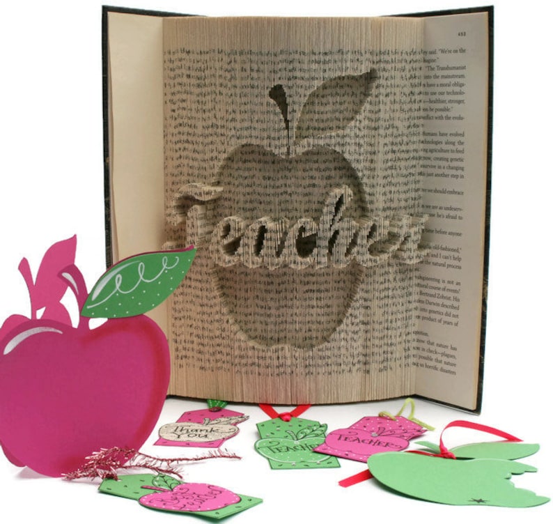 May include: A book with a red apple and the word "Teacher" cut out of the pages. The book is open and the pages are folded to create the shape of the apple. There are several small paper apple tags with the words "Thank You", "Teacher", and "Apple" on them.