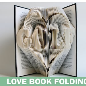 May include: A book with the pages folded into a heart shape, with the word "LOVE" folded into the centre of the heart. The book is open and the pages are folded in a way that creates a 3D effect. This is an example of book folding art.