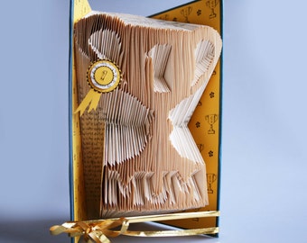 May include: A book folded into the shape of a trophy with a yellow ribbon and a gold medal with the number "1" on it. The book is standing on a yellow surface with a blue border and a pattern of small black cups.