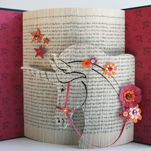 Peut inclure: Un livre en papier blanc plié en forme de tête de licorne. La licorne a un ruban rose pour bride et des fleurs en papier orange et rose sont attachées à la crinière. Il y a des étoiles en papier orange sur le fond.