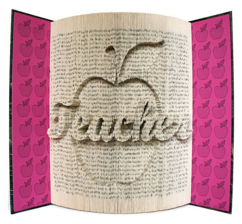 May include: A book with pages folded into the shape of an apple with the word "Teacher" inside. The cover of the book is pink with a pattern of red apples.