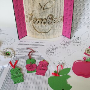 May include: A craft project for teachers with a book folded into the shape of an apple with the word "Teacher" written on it. There are several paper apple tags with different messages, including "Best Teacher", "Thank You", and "Teacher". There are also paper flower templates and a sheet of paper with a grid and the words "Apple Teacher" on it.
