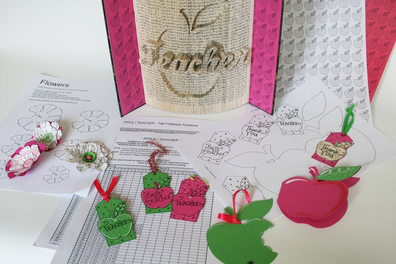 May include: A craft project for teachers with a book folded into the shape of an apple with the word "Teacher" written on it. There are several paper apple tags with different messages, including "Best Teacher", "Thank You", and "Teacher". There are also paper flower templates and a sheet of paper with a grid and the words "Apple Teacher" on it.