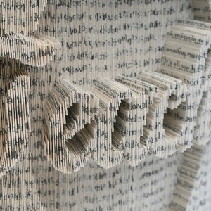 May include: A book art sculpture made from the pages of a book, folded and shaped to create the word "LOVE". The pages are white with black text.