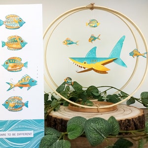 May include: A wooden hoop mobile with a blue and yellow shark, and several smaller yellow and blue fish. The mobile is hanging from a wooden hoop and is on a wooden base. The text "Dare to be different" is on a card in the background.