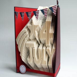 May include: A book art sculpture of the words "Happy 4th of July" with a red, white and blue bunting decoration. The sculpture is made from a book with pages folded into a three-dimensional design.