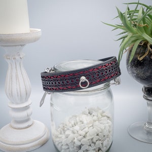 May include: A black leather collar with a red patterned design and a silver ring. The collar is sitting on top of a glass jar filled with white stones.
