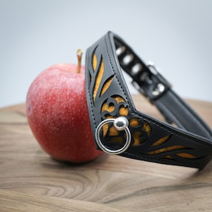 Könnte beinhalten: Ein schwarzes Lederhalsband mit goldfarbenem Blumendesign und einem silbernen Ring. Das Halsband liegt auf einer Holzoberfläche neben einem roten Apfel.