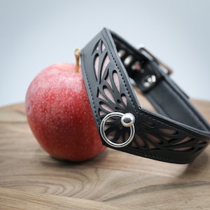 Könnte beinhalten: Ein schwarzes Lederhalsband mit einem rosafarbenen Blumendesign und einem silbernen Ring. Das Halsband liegt auf einer Holzoberfläche neben einem roten Apfel.