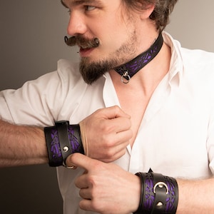 K&ouml;nnte beinhalten: Eine Person tr&auml;gt ein wei&szlig;es Hemd mit einem schwarzen Lederhalsband und passenden Armb&auml;ndern. Das Halsband und die Armb&auml;nder haben ein violettes geometrisches Muster.