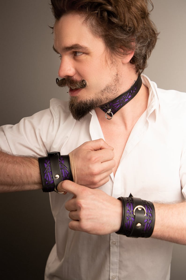 K&ouml;nnte beinhalten: Eine Person tr&auml;gt ein wei&szlig;es Hemd mit einem schwarzen Lederhalsband und passenden Armb&auml;ndern. Das Halsband und die Armb&auml;nder haben ein violettes geometrisches Muster.