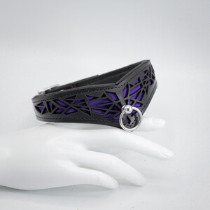 K&ouml;nnte beinhalten: Ein schwarzes Lederhalsband mit geometrischem Ausschnitt und einem violetten Futter. Das Halsband hat einen silbernen Metallring mit einer Kugel am Ende.