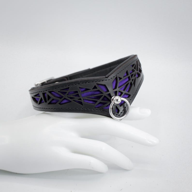 K&ouml;nnte beinhalten: Ein schwarzes Lederhalsband mit geometrischem Ausschnitt und einem violetten Futter. Das Halsband hat einen silbernen Metallring mit einer Kugel am Ende.
