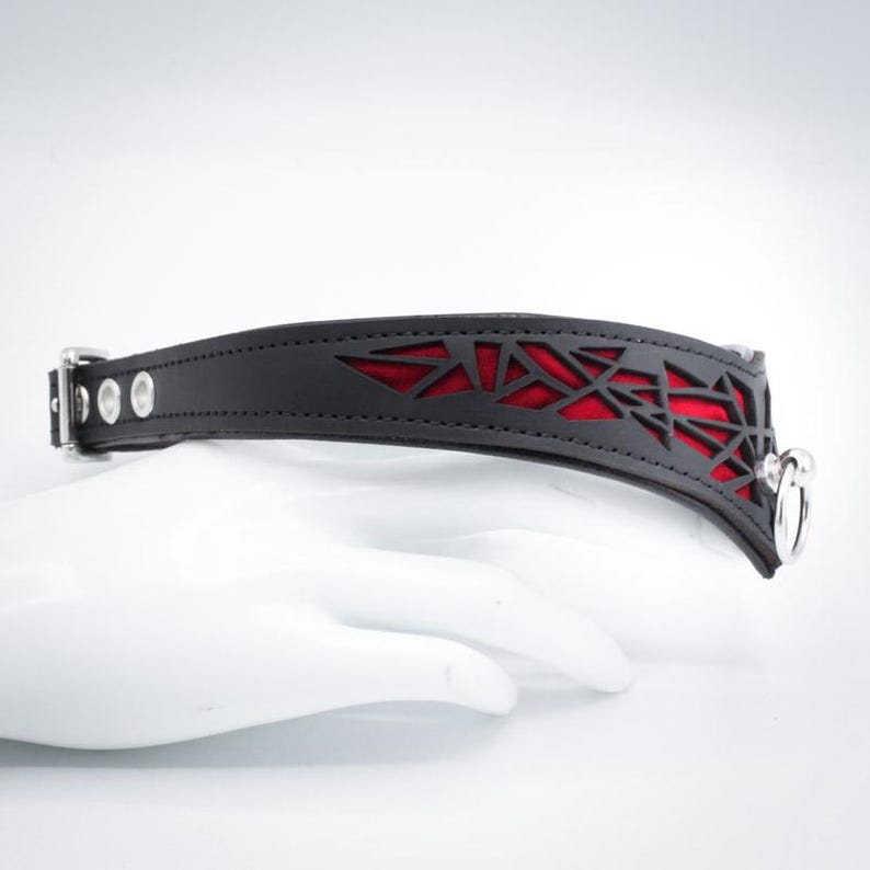 K&ouml;nnte beinhalten: Schwarzes Lederhalsband mit einem roten geometrischen Ausschnitt und silbernen Beschl&auml;gen.