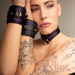 K&ouml;nnte beinhalten: Eine Person tr&auml;gt eine schwarze Lederkette mit einem violetten Spitzenmuster und dazu passende schwarze Lederhandgelenksmanschetten mit violetten Spitzenmustern. Die Person hat kurze Haare und T&auml;towierungen auf den Armen.
