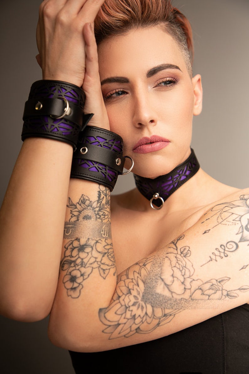 K&ouml;nnte beinhalten: Eine Person tr&auml;gt eine schwarze Lederkette mit einem violetten Spitzenmuster und dazu passende schwarze Lederhandgelenksmanschetten mit violetten Spitzenmustern. Die Person hat kurze Haare und T&auml;towierungen auf den Armen.
