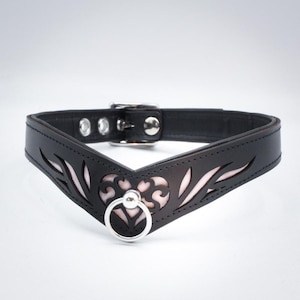 Könnte beinhalten: Schwarzes Lederhalsband mit dekorativem Ausschnittmuster und einem silberfarbenen Metallring. Das Halsband hat eine V-förmige Vorderseite mit hellrosa Innenfutter, das durch die Ausschnitte sichtbar ist. Es wird mit einer Schnalle befestigt.