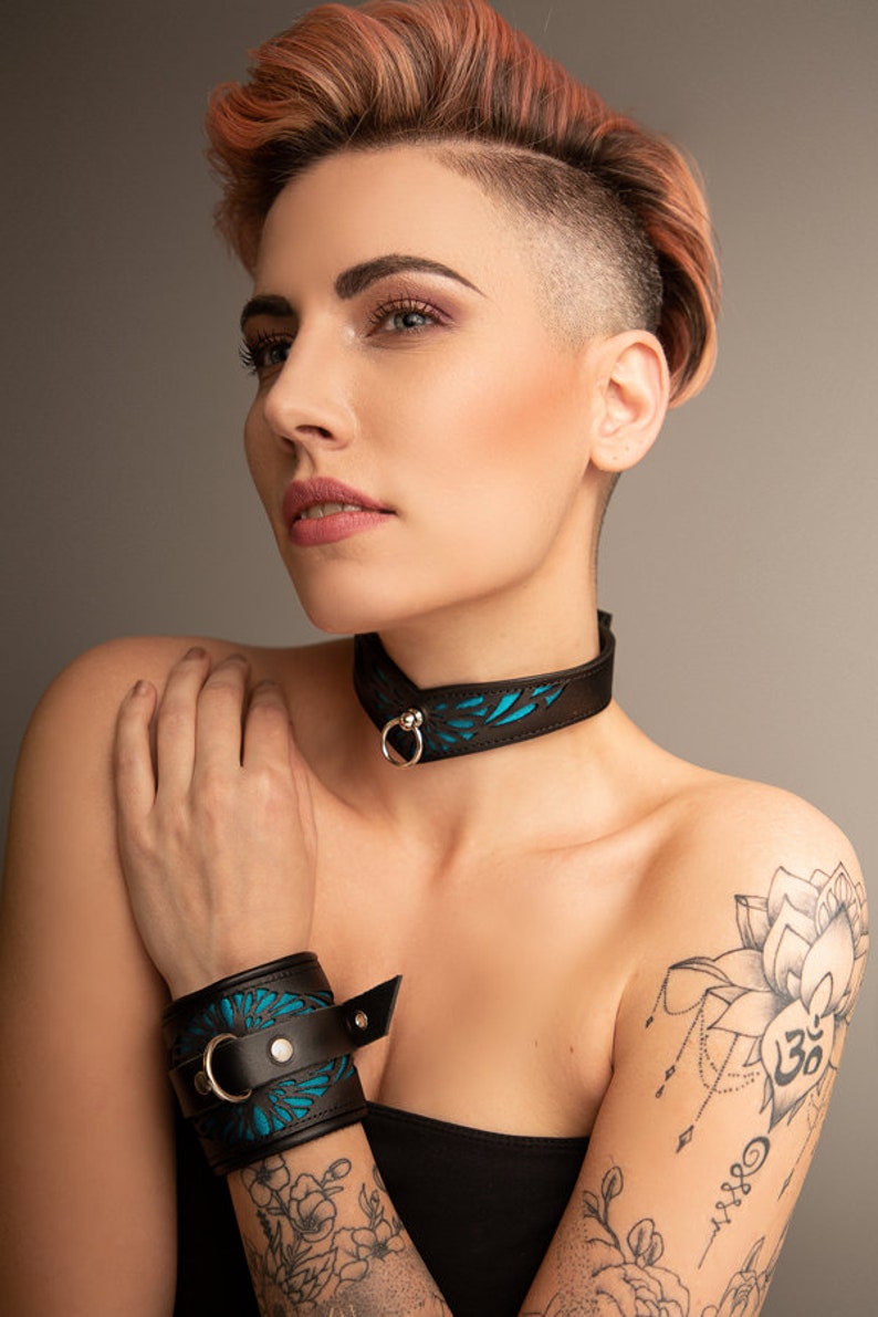 May include: A woman with a short, pink and blonde hairstyle is wearing a black leather choker with a blue floral design and a matching black leather wrist cuff. The choker and cuff have silver metal accents. The woman has a tattoo of a lotus flower and the word "Om" on her upper arm.
