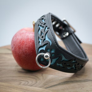 Könnte beinhalten: Schwarzes Lederhalsband mit blauem Blumendesign und einem silbernen Ring. Das Halsband liegt auf einer Holzoberfläche neben einem roten Apfel.