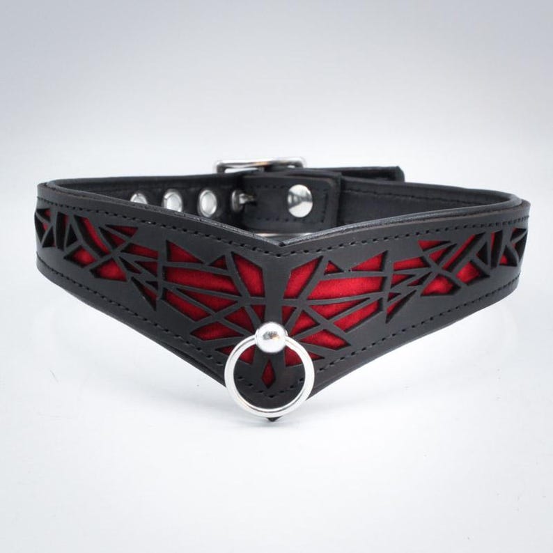 K&ouml;nnte beinhalten: Ein schwarzes Lederhalsband mit einem roten geometrischen Muster und einem silbernen Ring. Das Halsband ist mit Ausschnitten gestaltet, die das rote Stoff darunter freilegen.
