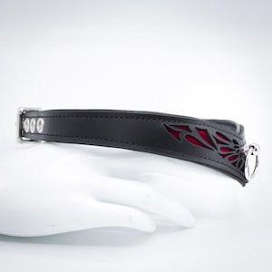 May include: Black leather collar with a red accent and a silver metal ring. The collar has a decorative cut-out design.