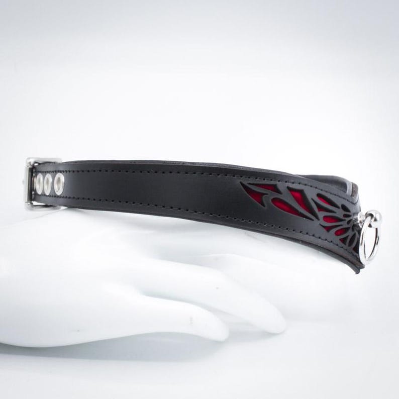 May include: Black leather collar with a red accent and a silver metal ring. The collar has a decorative cut-out design.
