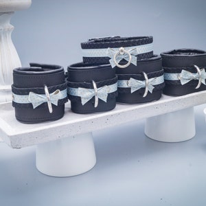 May include: A set of black leather cuffs and restraints with light blue patterned ribbon and silver-toned metal accents. The cuffs are displayed on a white pedestal tray, with a small cactus in a white pot to the side.