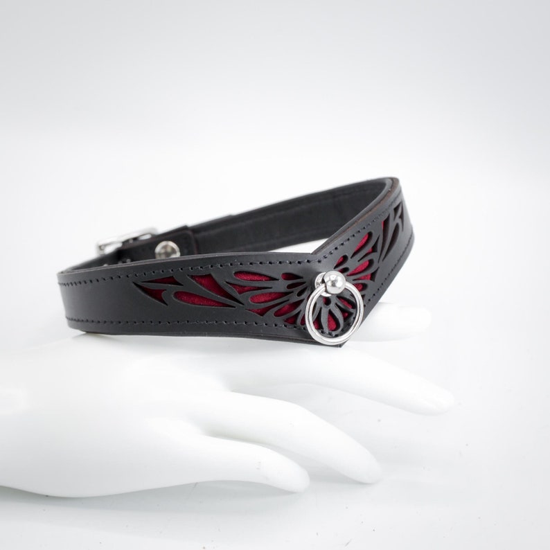 May include: Black leather collar with a red velvet inlay and a silver ring. The collar has a cut-out design of a stylised butterfly.