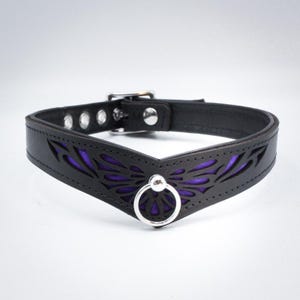 May include: A black leather choker with a purple butterfly design and a silver ring. The choker has a V-shaped front and is decorated with a cut-out butterfly pattern.
