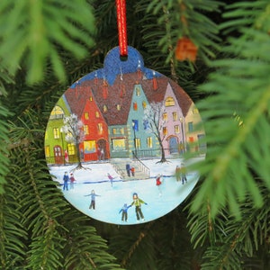 May include: A round Christmas ornament painted with a winter scene of a small town with houses, a frozen lake, and people ice skating. The ornament is hanging on a green pine tree branch.
