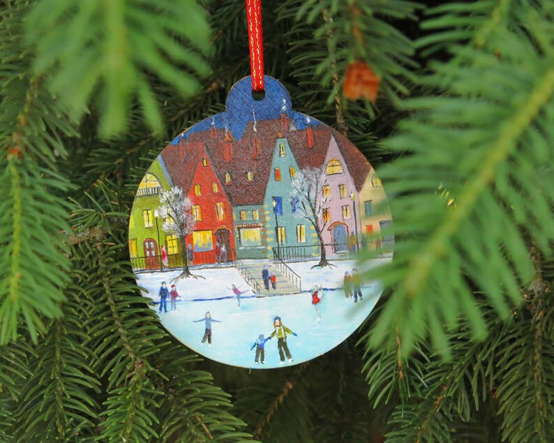 May include: A round Christmas ornament painted with a winter scene of a small town with houses, a frozen lake, and people ice skating. The ornament is hanging on a green pine tree branch.