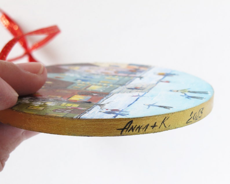 May include: A round wooden ornament with a painted scene of a winter village with people ice skating. The ornament has a gold rim and is signed "Anna + K. 2023".