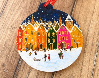 May include: A hand-painted wooden Christmas ornament depicting a snowy village scene with colorful houses, a church steeple, and a Christmas tree. The ornament is decorated with white snow and stars.