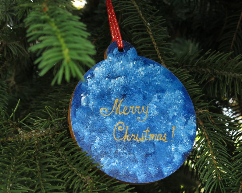 May include: Blue and white Christmas ornament with gold lettering that reads "Merry Christmas!" hanging on a green pine tree branch.