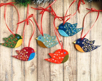 May include: Six wooden bird ornaments with painted floral patterns and blue, red, green, and yellow accents. The birds are hanging from red ribbon on a wood background.