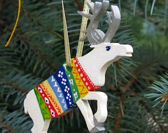 May include: A white wooden reindeer ornament featuring silver antlers and hooves, wearing a painted blanket with colorful horizontal stripes in green, red, yellow, and blue, suspended by a thin gold ribbon for hanging on a tree branch.