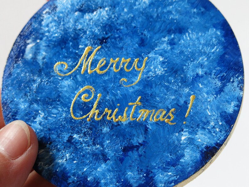 May include: Round wooden ornament painted in a blue and white marbled pattern with the words "Merry Christmas!" written in gold cursive.