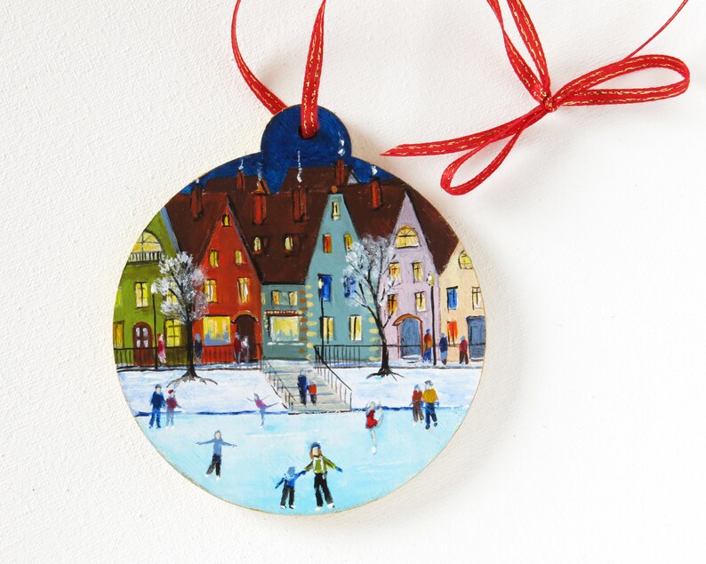 May include: A hand-painted wooden ornament depicting a winter scene with colorful houses, a frozen lake, and people ice skating. The ornament is round and has a red ribbon attached.