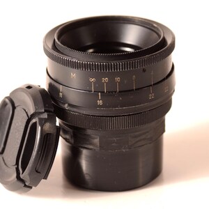May include: A black camera lens with a detached lens cap. The lens has markings for various settings, including numbers and symbols. The lens cap is also black and sits to the left of the lens.