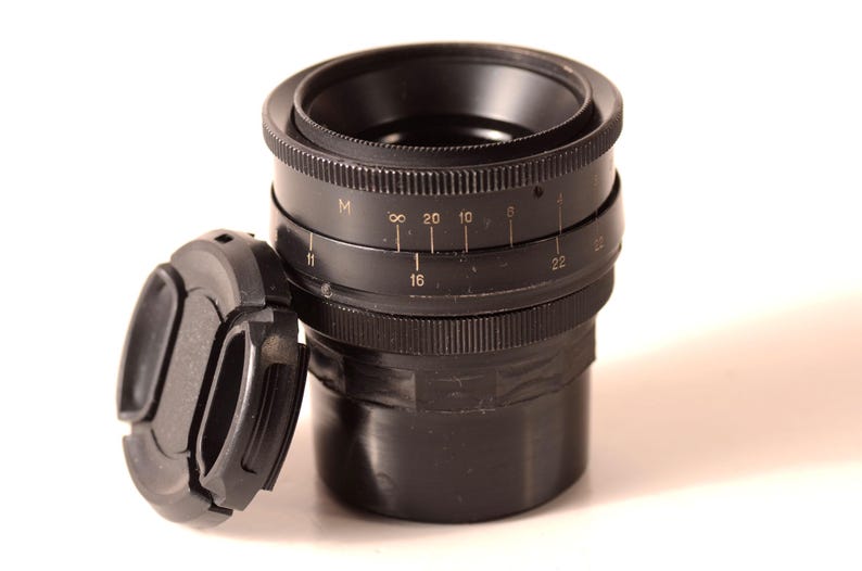 May include: A black camera lens with a detached lens cap. The lens has markings for various settings, including numbers and symbols. The lens cap is also black and sits to the left of the lens.