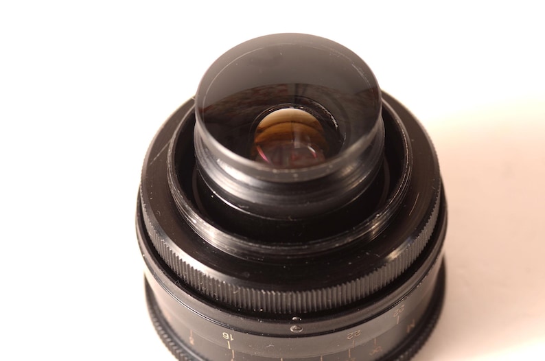 May include: Close-up of a black camera lens with a clear, rounded lens element. The lens has concentric rings and a ridged focus ring. The lens is set against a white background.