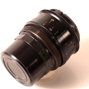 May include: A black camera lens with a ribbed focus ring and aperture markings. The lens has a cylindrical body and is angled slightly, showcasing its various settings and measurements. The lens is set against a white background.