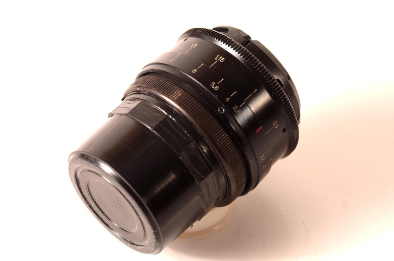 May include: A black camera lens with a ribbed focus ring and aperture markings. The lens has a cylindrical body and is angled slightly, showcasing its various settings and measurements. The lens is set against a white background.
