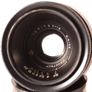 May include: A black Jupiter R-12 2.8/55 camera lens. The lens features a circular design with a central aperture and markings including "JUPITER R-12", "2.8/55", and "N8407307". The lens has a series of aperture settings.