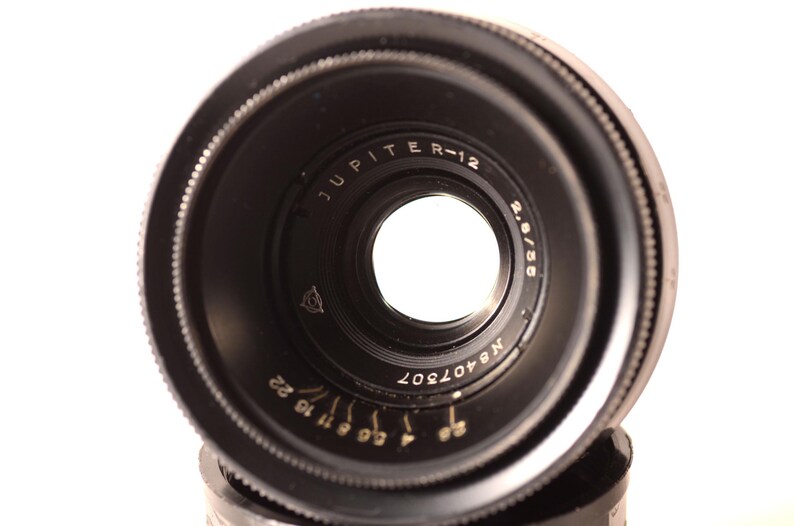 May include: A black Jupiter R-12 2.8/55 camera lens. The lens features a circular design with a central aperture and markings including "JUPITER R-12", "2.8/55", and "N8407307". The lens has a series of aperture settings.