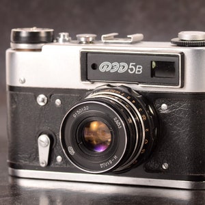 May include: A silver and black 35mm film camera with a black leather covering. The camera has a lens with a black ring and a silver inscription that reads "ФЭД 5B".
