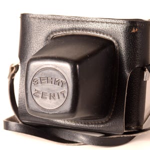 May include: Black leather camera case with a shoulder strap. The case has a square, textured design with a circular emblem that reads "ZENIT". The strap is a light grey colour.