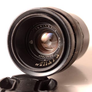 May include: A black Jupiter-12 camera lens with a lens cap. The lens has a series of numbers and markings, including "Jupiter-12" and "f/2.8 50". The lens is angled towards the viewer, with the lens cap in the foreground.