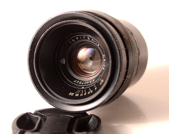 May include: A black Jupiter-12 camera lens with a lens cap. The lens has a series of numbers and markings, including "Jupiter-12" and "f/2.8 50". The lens is angled towards the viewer, with the lens cap in the foreground.
