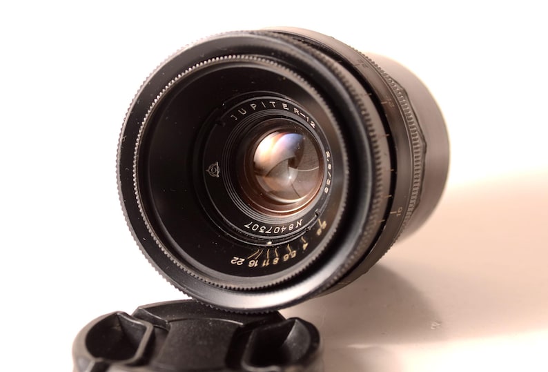 May include: A black Jupiter-12 camera lens with a lens cap. The lens has a series of numbers and markings, including "Jupiter-12" and "f/2.8 50". The lens is angled towards the viewer, with the lens cap in the foreground.